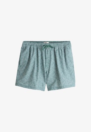 Homme torse nu portant des lunettes de soleil et un short de bain se tient pieds nus sur un rocher près de la plage, tenant une serviette beige autour du cou.