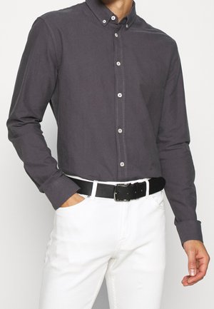 Mężczyzna w ciemnoszarej koszuli zapinanej na guziki, wsadzonej w białe spodnie z czarnym paskiem, jedną ręką w kieszeni, na jednolitym jasnym tle.