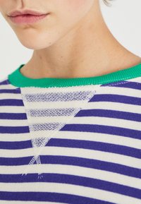 Maglione a righe con strisce orizzontali bianche e viola, con un collo a coste verde brillante e un motivo triangolare testurizzato sul petto.