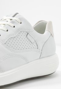 Białe skórzane trampki z perforowanym panelem bocznym, białymi sznurówkami, wyściełaną podeszwą i miękkim beżowym detalem przy kołnierzu pięty.
