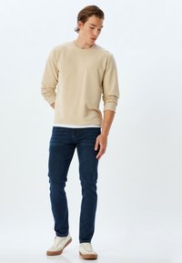 Sudadera beige con cuello redondo y puños acanalados, combinada con jeans slim-fit de color azul oscuro. Zapatillas blancas con suelas beige completan el conjunto.