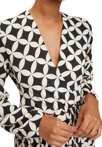Černé šaty s bílým kulatým vzorem, s hlubokým výstřihem do V, dlouhými rukávy, elastickým pasem a nařasenými manžetami. Měkká, texturovaná tkanina.