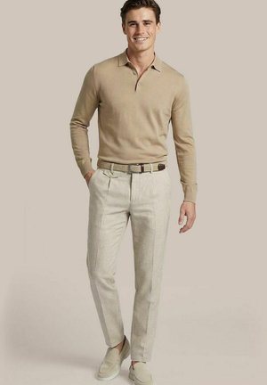 Jonge man die lacht, draagt een beige poloshirt met lange mouwen, lichtgekleurde broek met geweven riem en lichte instapschoenen, staand voor een effen achtergrond.