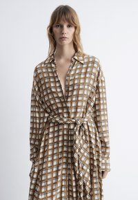 Langärmliges, braun kariertes Kleid mit Taillengürtel, Kragen und knöpfbarem Vorderteil. Der Stoff hat eine glatte Textur.