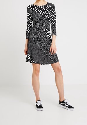 Kobieta ubrana w czarno-białą sukienkę do kolan w mieszany wzór w kropki, z rękawami do łokcia, oraz czarne trampki z białymi sznurowadłami.
