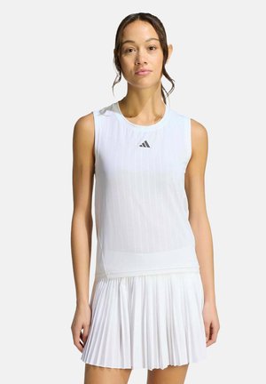 Vrouw draagt witte mouwloze Adidas top en witte geplooide rok, staand tegen een effen lichte achtergrond.