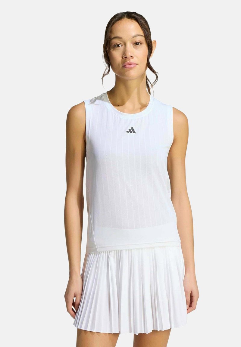 Femme portant un haut blanc sans manches Adidas et une jupe blanche plissée, debout devant un fond clair uni.