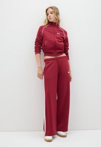Chándal burdeos con detalles de ribete blanco, que presenta una chaqueta corta con cremallera y pantalones de pierna ancha, fabricado en un tejido suave.