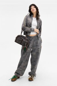 Šedá bunda se vzorem camouflage na zip s odpovídajícími tepláky. Hnědá kabelka se dvěma zipovými kapsami a zeleným klíčenkou jako akcentem. Hnědé boty.