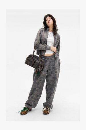Šedá bunda se vzorem camouflage na zip s odpovídajícími tepláky. Hnědá kabelka se dvěma zipovými kapsami a zeleným klíčenkou jako akcentem. Hnědé boty.