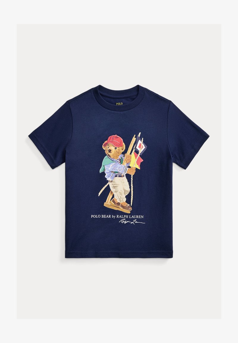 Camiseta para niño pequeño azul marino con un gráfico de un oso con una gorra roja sosteniendo banderas náuticas, una cuerda y vistiendo ropa casual.