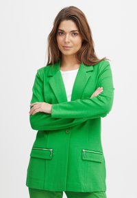 Blazer verde de tejido texturizado, que presenta dos bolsillos frontales con detalles de cremallera plateada y cierre de un solo botón.
