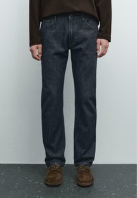 Mann trägt dunkelblaue Gerade-Passform-Jeans, braune Wildleder-Schnürschuhe und ein dunkles Langarmshirt, steht auf einem gefleckten Boden.