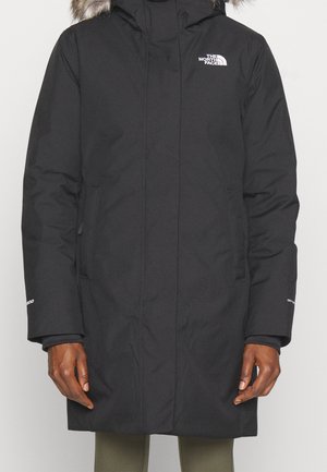 Persoon die een lange zwarte North Face winterjas met een capuchon met bontkraag draagt, staand met handen ontspannen langs het lichaam.