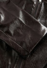 Détail d'une veste en cuir synthétique noire avec une texture brillante, une fermeture éclair visible et une finition froissée. Mise au point sur la zone de la manche et la qualité du tissu.