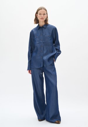 Denim shirt with long sleeves, two chest pockets, and button closure. Paired with wide-leg denim trousers and animal print shoes. Blue hues throughout.