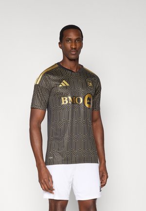 Black and gold soccer jersey with geometric pattern, short sleeves, rounded neck, and branding in gold. Worn with white shorts.