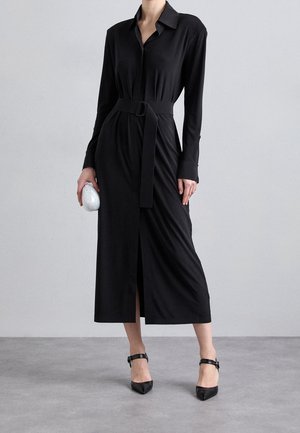 Kvinde iført en lang sort skjortekjole med bælte og lange ærmer, der holder en lille sølvfarvet clutch og sorte ankelrem-sandaler med hæl.