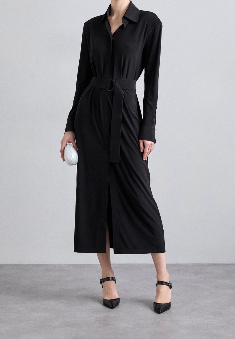 Femme portant une longue robe chemise noire ceinturée à manches longues, tenant une petite pochette argentée et des talons noirs à bride à la cheville.