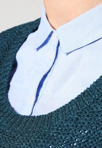 Zbliżenie na jasnoniebieską koszulę z guzikami, której kołnierz wystaje spod ciemnozielonego, dzianego swetra z teksturowanym wzorem.