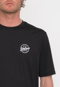 Camiseta negra de manga corta con cuello redondo, fabricada en tela ligera. Presenta un logo blanco de "Volcom" en el área del pecho izquierdo.