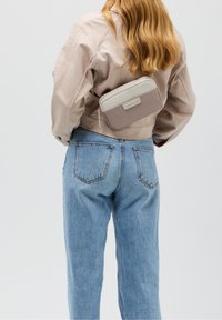 Borsone beige realizzato in tessuto liscio con un accento grigio chiaro, caratterizzato da una zip e una toppa con logo, posizionato sulla schiena di una persona.