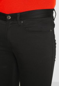 Primo piano di jeans neri con bottone e cuciture argentate, indossati con una maglia rossa brillante.