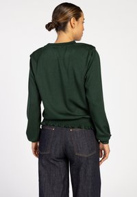 Donkergroene pullover met een geplooide, textuurachtigezoom en lange mouwen, gecombineerd met donkerdenim jeans. Glad materiaal met minimale details.