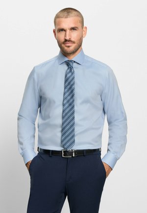 Man met kort haar en baard, die een lichtblauw overhemd, gestreepte blauwe das, donkerblauwe broek en zwarte riem draagt, handen in de zakken.