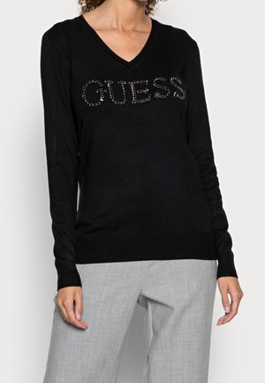Pullover nero a collo a V realizzato in tessuto morbido, con logo 'GUESS' impreziosito da strass sul davanti, abbinato a pantaloni grigi.