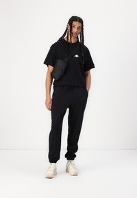 Zwarte oversized t-shirt met logo, zwarte joggingsbroek met koord, en beige sneakers. Inclusief een kleine zwarte schoudertas. Effen achtergrond.