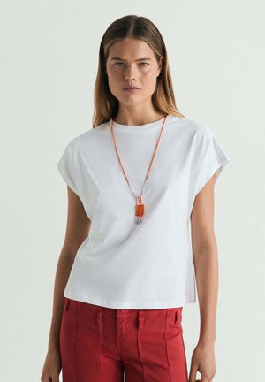Mujer con cabello liso lleva una camiseta blanca sin mangas, pantalones rojos y un collar naranja de cuentas sobre un fondo claro y liso.