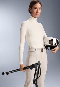 Hladký biely lyžiarsky outfit s vysokým golierom, kombinovaný so svetlými béžovými nohavicami, čiernymi lyžiarskymi palicami a bielou prilbou s clonou.