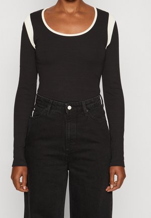 Kvinde iført en sort langærmet top med hvide kantdetaljer på skuldre og halsudskæring, kombineret med sorte højhængende jeans.