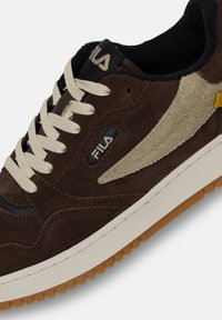 Bruna mockaskor med beige accenter, som har krämfärgade snören, svart foder och en gummisul med ett strukturerat mönster. Fila-logotypen är synlig.