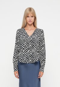 Blouse à imprimé zèbre noir et blanc avec un décolleté en V, des manches longues et une coupe décontractée. Portée par-dessus une jupe unie bleu marine.
