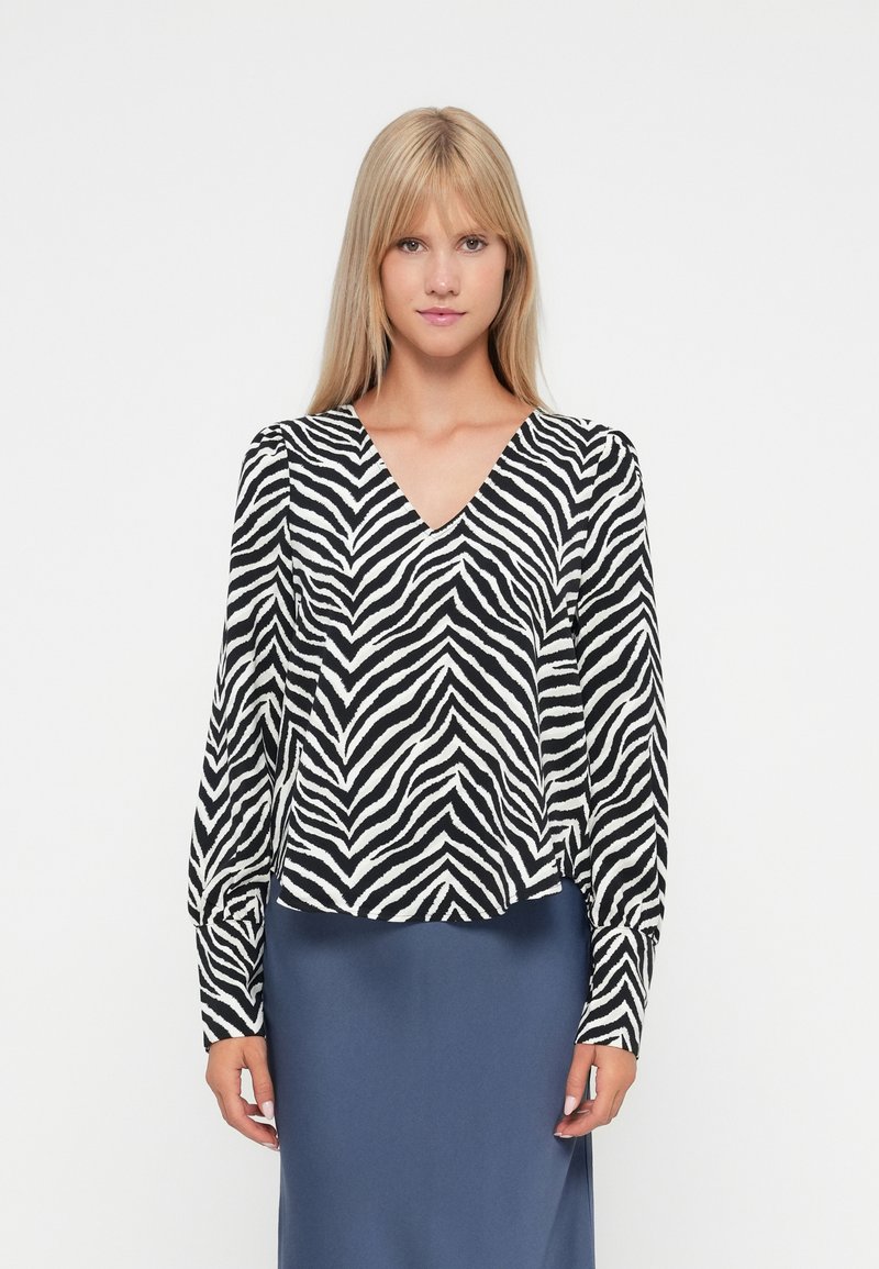 Blouse à imprimé zèbre noir et blanc avec un décolleté en V, des manches longues et une coupe décontractée. Portée par-dessus une jupe unie bleu marine.