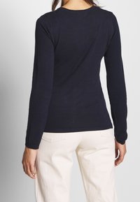 Kvinna i en figurnära marinblå långärmad topp och högmidjade off-white byxor, sedd bakifrån mot en enfärgad bakgrund.