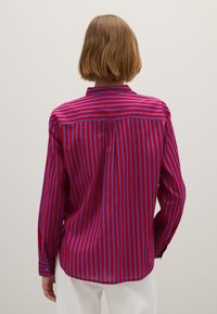 Persoon met kort lichtbruin haar staat met zijn gezicht naar een muur, gekleed in een rood en blauw verticaal gestreept long-sleeve shirt en een witte broek.