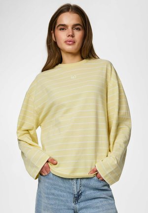Ung kvinde med langt brunt hår iført en løs lysegul striped langærmet skjorte og blå jeans, hænder i lommerne.