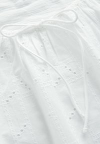 Tissu blanc avec motifs géométriques brodés et petits trous, doté d'un cordon noué en boucle en haut.