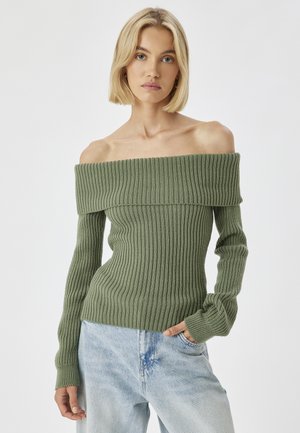 Ein off-shoulder, grüner, gerippter Pullover mit langen Ärmeln, aus einem strukturierten Strick gefertigt, kombiniert mit hellblauen Denim-Jeans.