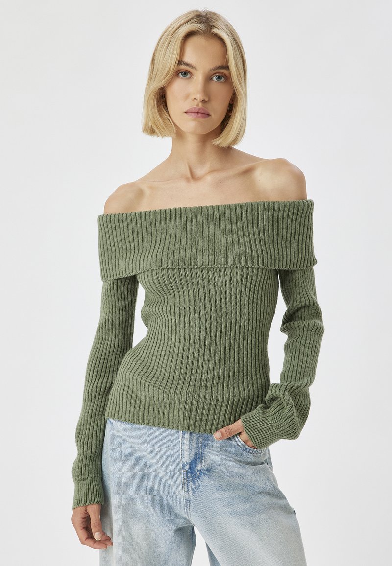 Ein off-shoulder, grüner, gerippter Pullover mit langen Ärmeln, aus einem strukturierten Strick gefertigt, kombiniert mit hellblauen Denim-Jeans.