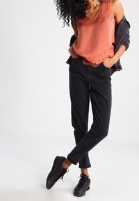 Czarne zwężane spodnie z podwiniętymi mankietami, w połączeniu z luźnym bezrękawnikiem w kolorze rdzy, noszonym pod ciemną kurtką oraz czarnymi sznurowanymi butami.