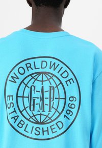 Világoskék póló fekete grafikával, rajta a "GAP" felirattal és egy földgömb mintával. A szöveg tartalmazza a "WORLDWIDE" és az "ESTABLISHED 1969" feliratokat. Puha anyag.