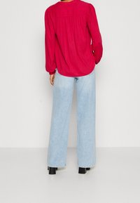 Röd blus med långa ärmar och rynkade axeldetaljer, matchad med ljusblå vida jeans och svarta ankelboots. Minimalistisk design.