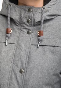 Šedá prošívaná bunda s kapucí, kovovými patentkami a stahovacími šňůrkami s hnědými koženými akcenty a textura povrchu.