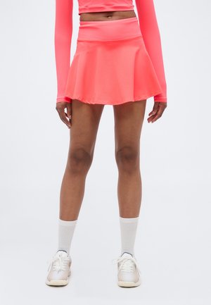 Moteris dėvinti koralų spalvos sportinę sijoną ir viršų, baltas vidutinio ilgio kojines ir baltus sportinius batelius, stovinti priešais paprastą foną.