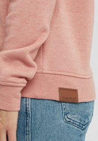 Felpa rosa chiaro in tessuto testurizzato; presenta un'etichetta in pelle con "oxmo" sull'orlo, abbinata a jeans in denim blu.