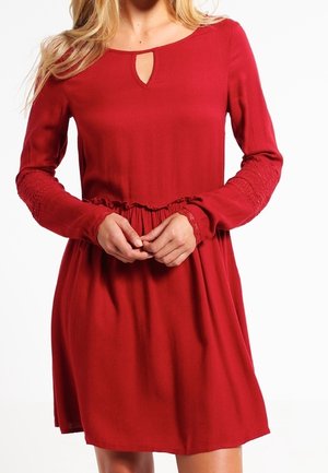 Kvinde iført en rød kjole med lange ærmer, der går til knæene, med en lille nøglehulskæring ved halsen og blonde-detaljer på ærmerne.